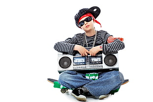 boy sitting on skateboard while carrying a cassette radio on lap