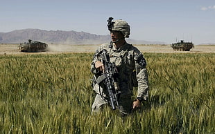 soldier holding black assault rifle standing between green grass field behind 2 battle tanks during daytime