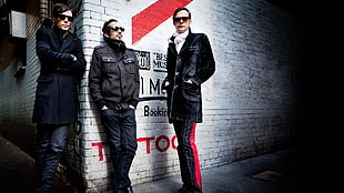 three men in black leather jacket leaning on white concrete wall