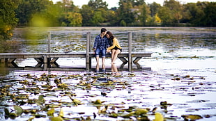 man and woman both sitting on gray wooden dock on lake under blue sky at daytime
