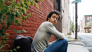 man wears gray long-sleeved shirt and blue denim jeans beside brown concrete wall during daytime