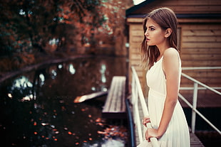 woman in white sleeveless dress standing on bridge near house and calm body of water during daytime