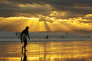 man carrying surfboard beside sea during sunset