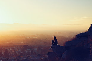landscape silhouette photography of person sitting on cliff facing urban area during golden hour