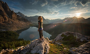 woman standing on rock ledge facing lake between trees under cloudy sky
