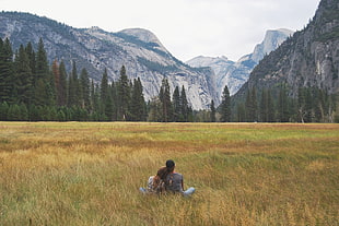 two person sitting in grass field while facing the wilderness