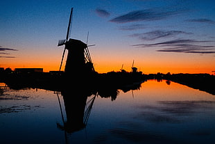 silhouette photo of windmill beside body of water during golden hour
