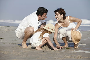 photo of man , woman and child playing sand at sea shore while smiling