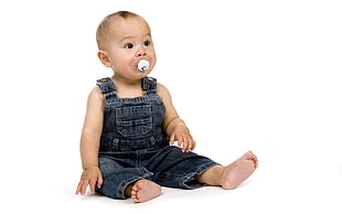baby with pacifier and wearing blue overalls laying on white floor