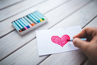 person holding pink crayon near pink heart shape sketch