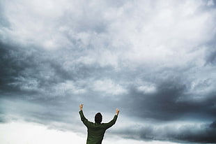 man wearing green dress shirt standing and raising hands under grey clouds at daytime