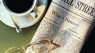 photo of white ceramic mug full with black coffee near Wall Street newspaper and gold-colored frame eyeglasses