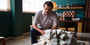 man wearing white button-up shirt sitting on white wooden armchair near pile of bundle of US dollar bills