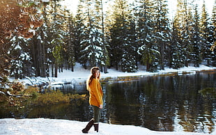 woman in orange jacket and black knee-high boots standing beside body of water