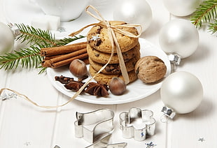 stack of cookies near cinnamon sticks, hazel nuts, and star anise on white ceramic plate near white bauble decor