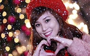 smiling woman wearing knit cap forming heart gesture with hands and lights in background