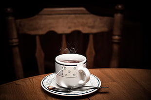 white-and-black floral print teacup and saucer filled with black coffee on brown wooden table near brown wooden bannister chair