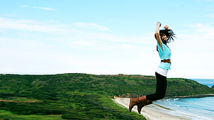 women in teal shirt with black pants jumping towards ocean taken at daytime