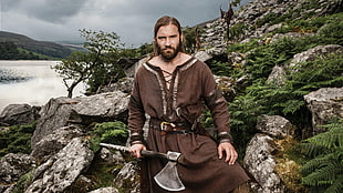 man standing on the side of the hill wearing brown robe holding axe during daytime