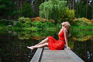 woman wearing red sundress sitting on gray wooden lake pier