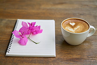 pink bougainvillea flower on white spiral notebook beside white ceramic up of latte