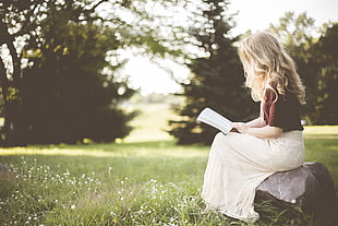 blonde haired woman sitting on rock while reading book during daytime