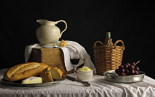 bread near white ceramic pitcher, brown basket with bottle and grape
