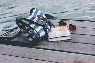 blue, white, and black stripe crossbody bag beside silver-framed aviator style sunglasses and book on brown wooden dock