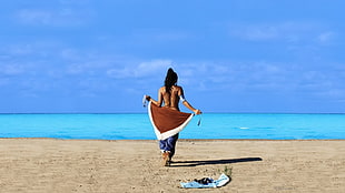 woman walking on shoreline holding scarf during daytime