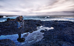man standing on a low-tide sea rocks while capturing photo using camera on tripod