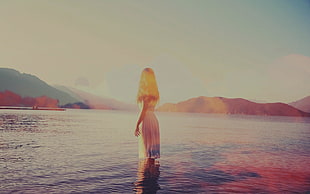 photo of woman wearing dress standing in body of water near mountains during day time