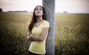 woman in yellow scoop-neck shirt leaning on brown wooden pole beside a white and yellow flower field