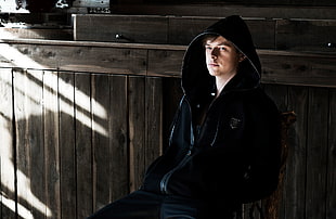 man wears black hooded jacket sits on wooden chair inside a well-lit room HD wallpaper