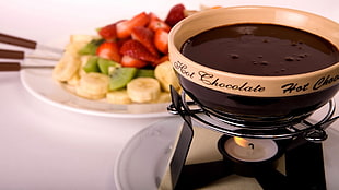 brown and black ceramic bowl filled with hot chocolate near sliced banana, kiwi, and strawberry fruits on white ceramic plate