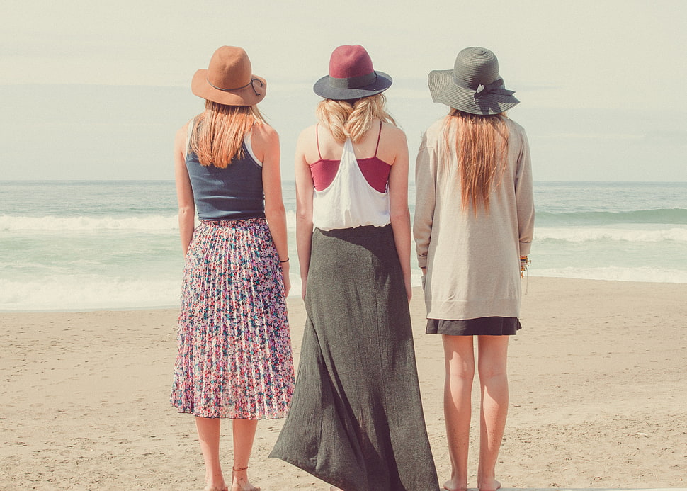 back view of three women in dress standing on seashore HD wallpaper