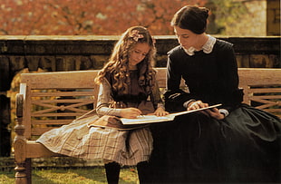 woman beside black long sleeve dress beside girl in brown long sleeve dress holding book sitting in brown wooden bench