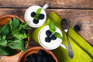 green leaves on brown wooden bowl beside two white ceramic mugs filled with white liquid and black berries on top placed on brown wooden panel