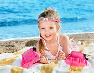 girl in pink swimwear and goggles lying down on sand in the seashore during daytime