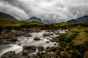 green grass field with river under cloudy sky with mountain background, sligachan