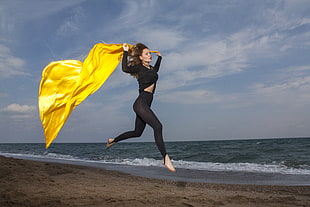 woman in black sweater and leggings holding yellow shawl jumping near seashore at daytime HD wallpaper