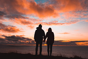 silhouette of man and woman standing on grass near ocean during golden hour