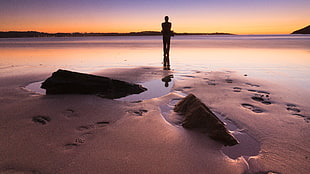 silhouette photography of person standing on beach sand during low tide and golden hour