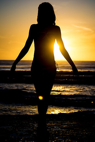 silhouette of woman standing on sea shore during sunset HD wallpaper
