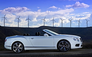 white Bentley convertible park on concrete road with silhouette of wind turbines