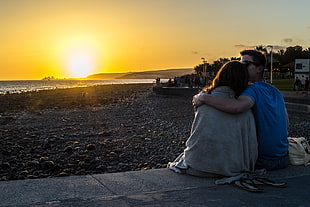 couple holding each other sitting near beach during golden hour HD wallpaper