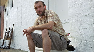 male wearing beige and gray collared shirt and gray cargo shorts sitting on white concrete surface