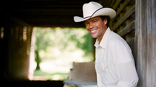 man wearing white cowboy hat and white dress shirt leaning on brown wooden wal