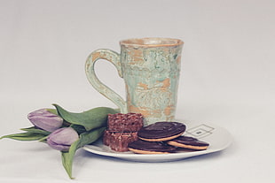 gray and beige ceramic mug on white ceramic plate with cookies and purple tulips