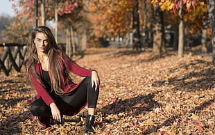 selective focus photography of woman in red cardigan kneeling on ground filled with dried leaves