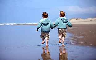 two children wearing teal hoodie jacket and beige shorts running on seashore